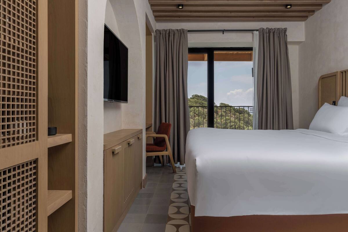 Una habitación de hotel con cama, televisor, silla y balcón con cortinas.