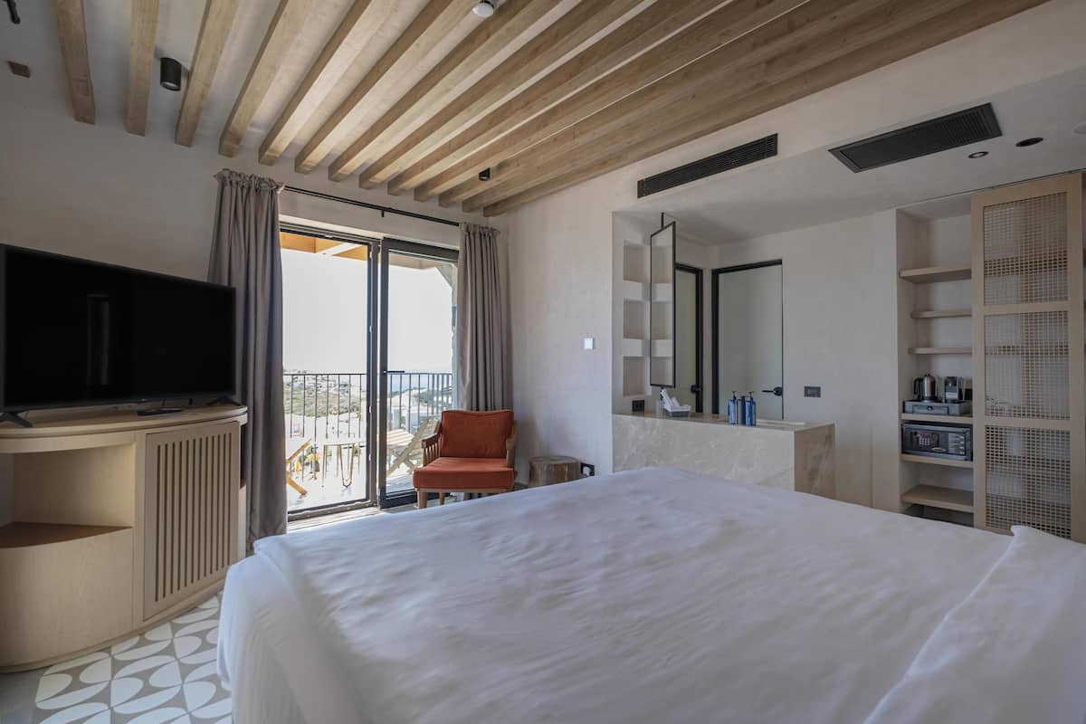 Habitación de hotel con una cama grande, televisión, una silla, balcón con vistas y baño con espejo y estantes.