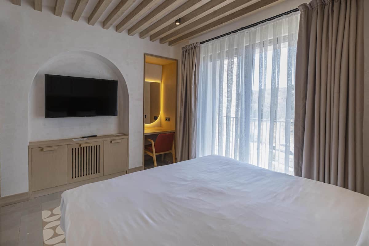 Habitación de hotel con una cama grande, un televisor en la pared, un escritorio con una silla y una ventana con cortinas.
