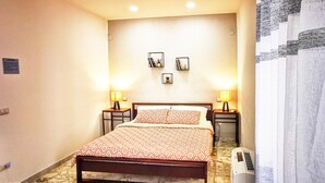 Une chambre d’hôtel comprenant un lit, deux tables de chevet avec des lampes, une étagère murale avec des livres et une plante, ainsi qu’un appareil de climatisation.