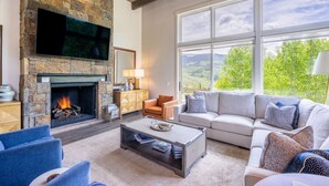 A spacious living room with a stone fireplace, a large window offering a view of greenery, a comfortable sofa set, and a flat-screen TV mounted above the fireplace. - Ultra Luxury Mountain Retreat | Private Hot Tub + Firepits | Privacy | Summit Sky Ranch (Silverthorne)