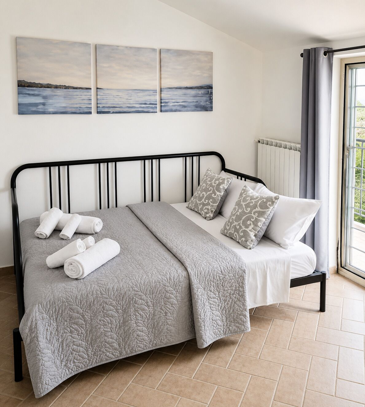 Una cama con colcha gris, almohadas blancas y toallas enrolladas. Hay tres cuadros colgados en la pared sobre la cama.