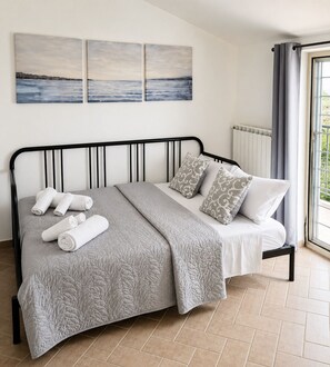 A bed with a grey quilt, white pillows, and rolled towels. There are three wall-mounted paintings above the bed. - To complete (Anguillara Sabazia)