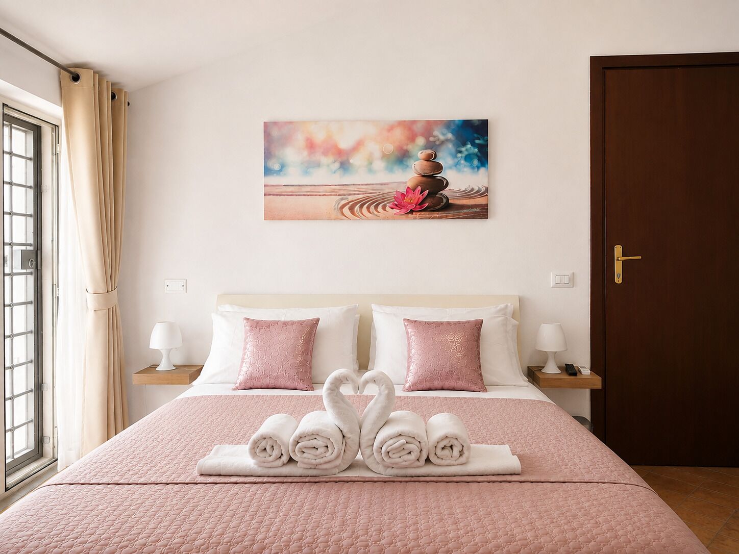 Una camera d'albergo con un letto, cuscini, un quadro, una porta e una finestra con tende.