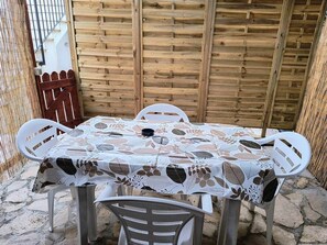 A round table with a floral tablecloth, surrounded by white plastic chairs, set against a wooden fence.