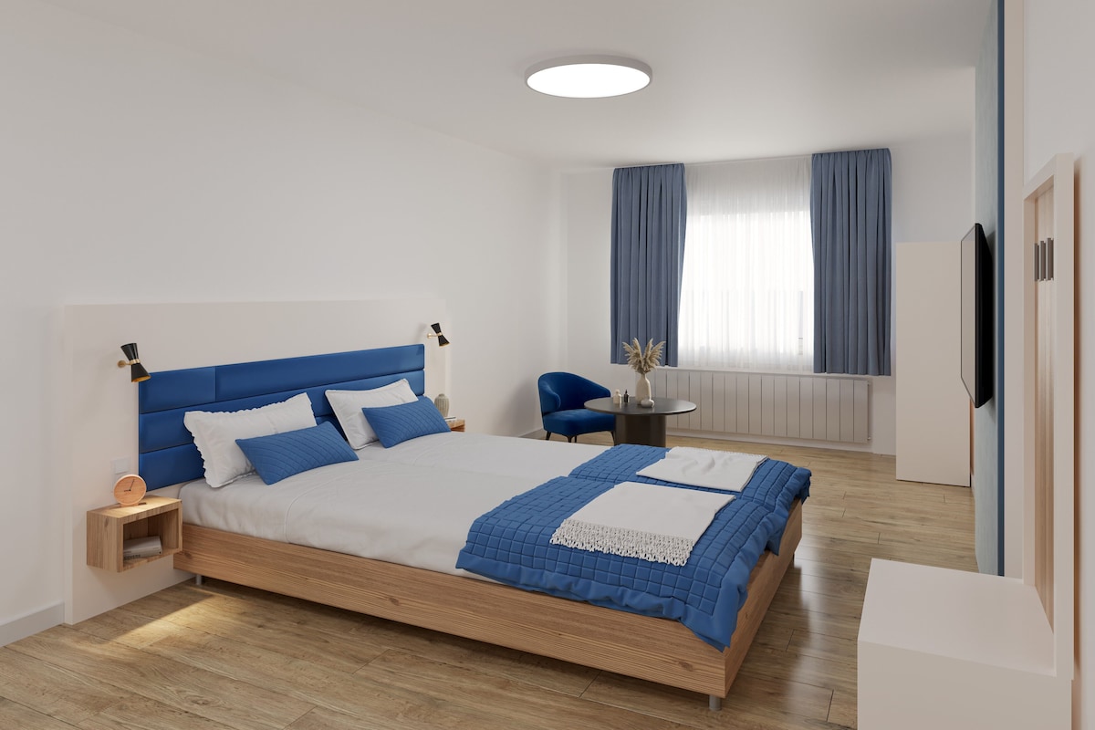 Un dormitorio moderno con cama de madera, mesita de noche, una silla, una mesa redonda y televisor montado en la pared.