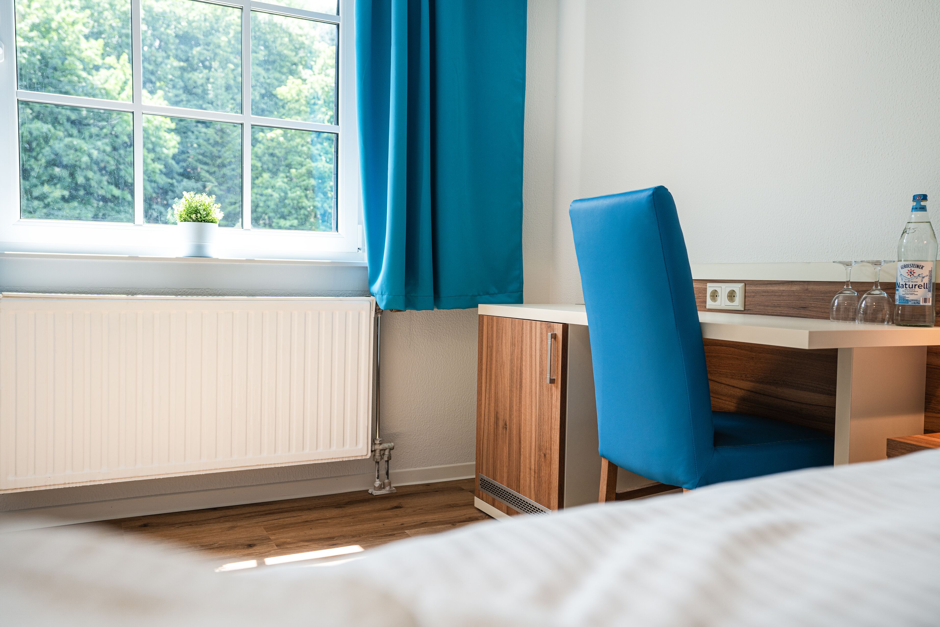 Een moderne hotelkamer met een houten vloer, een bed, een bureau met stoel, een raam met gordijnen en een radiator.