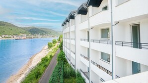 A multi-story hotel with balconies overlooking a river, a path along the riverbank, and a view of a town in the distance. - besttime Hotel Boppard (Boppard)
