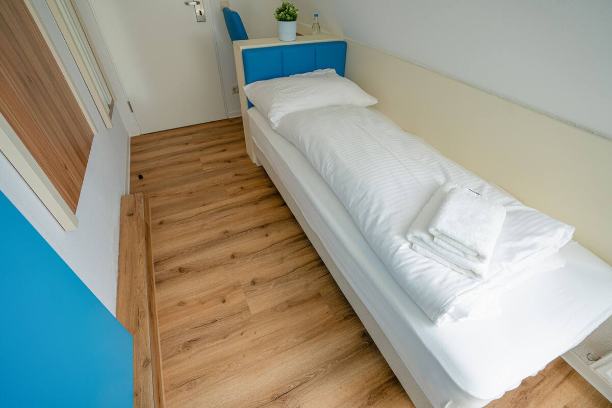 Un lit simple avec une literie blanche, une tête de lit bleue et une petite plante en pot sur la table de chevet.