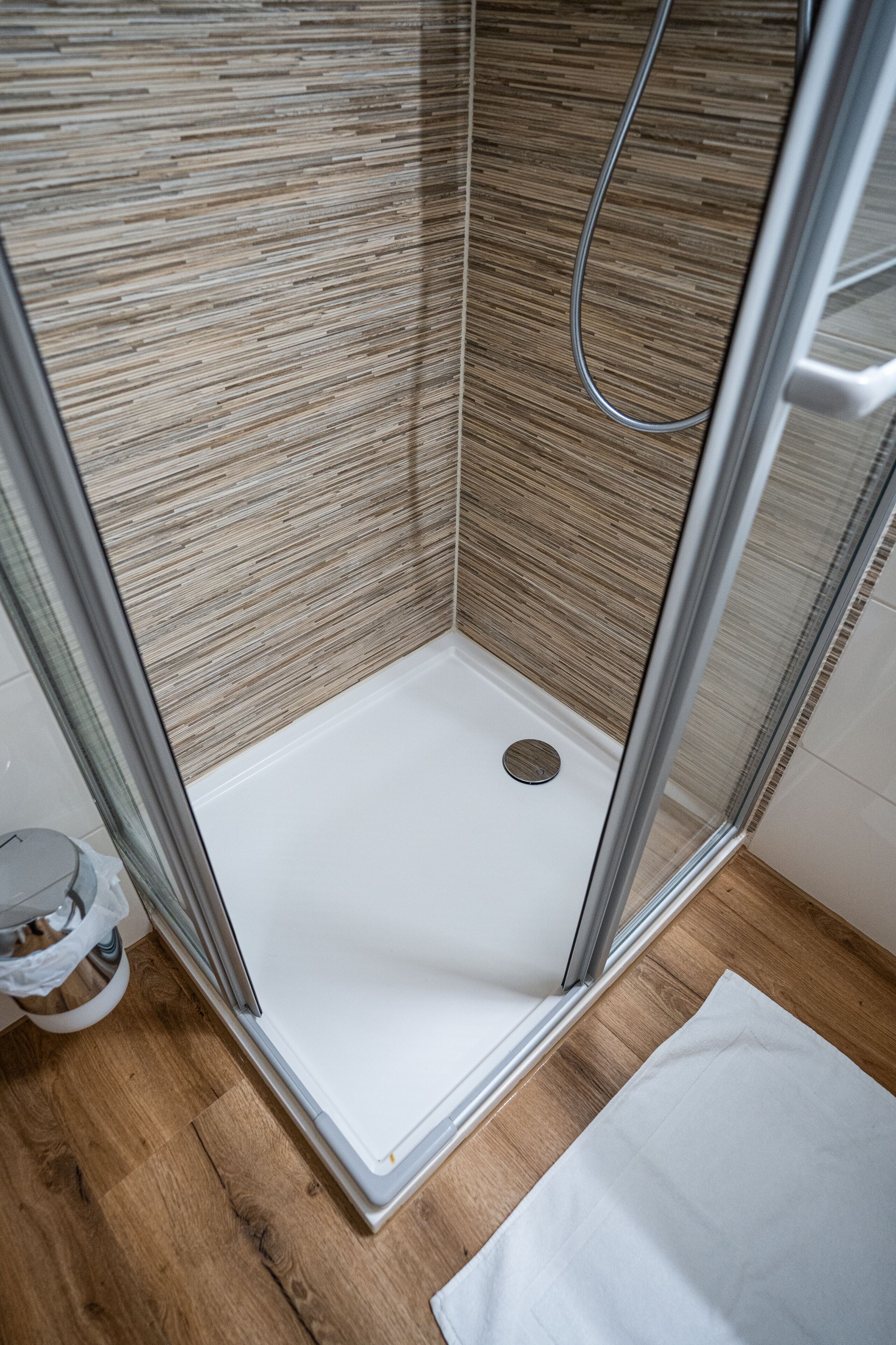 A modern shower with a glass door, a wooden floor, and a white mat.
