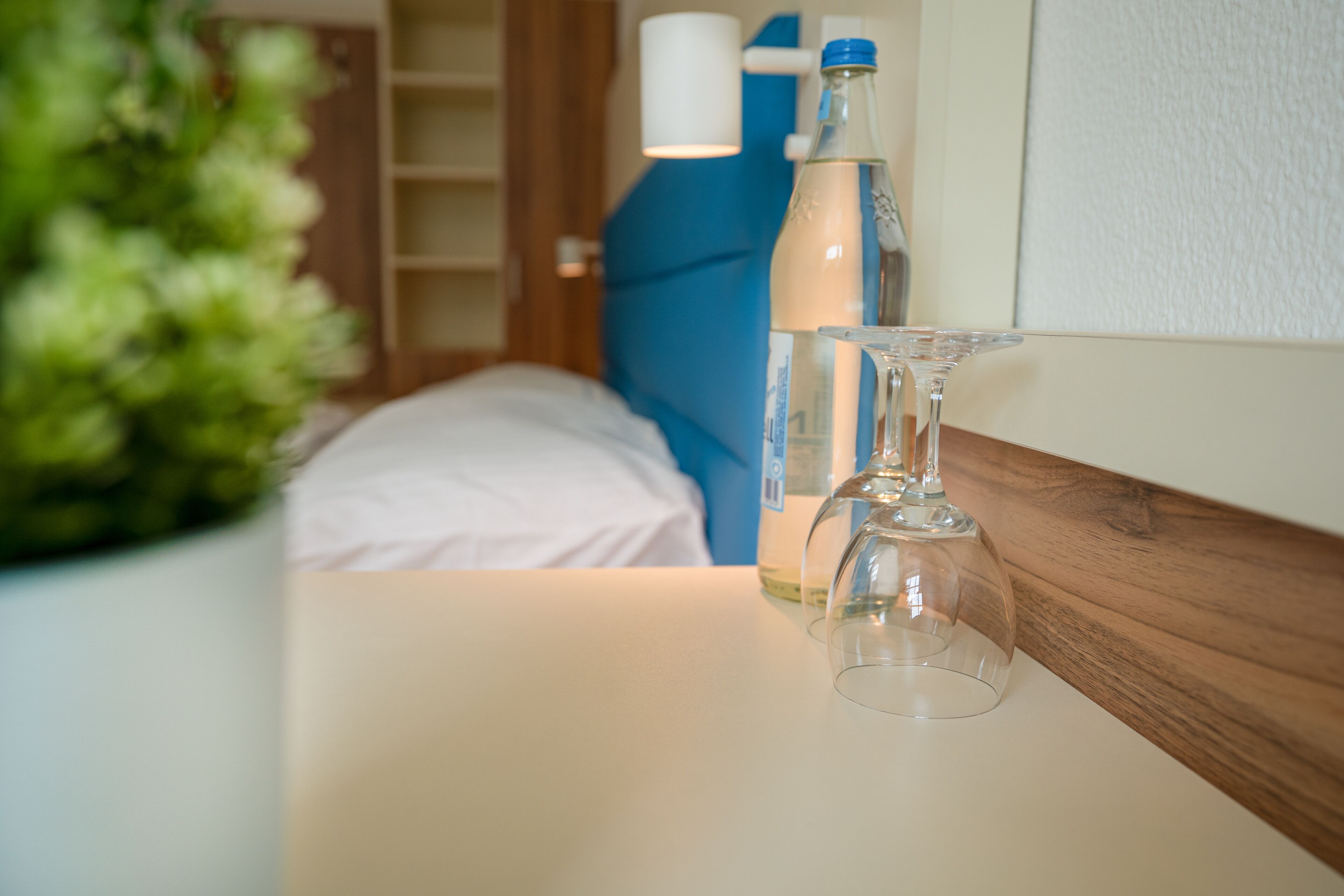 A hotel room with a bed, a bedside table with a bottle and a glass, and a potted plant.