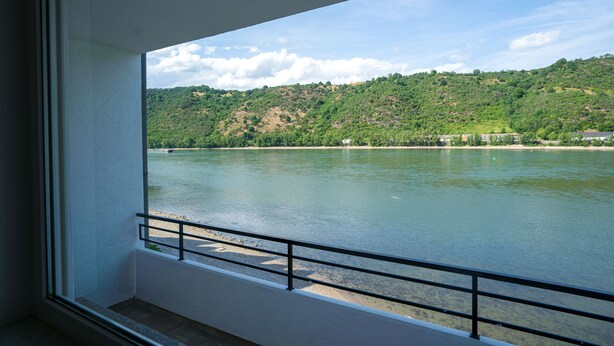 A room with a large window overlooking a river and a hilly landscape. - besttime Hotel Boppard (Boppard)