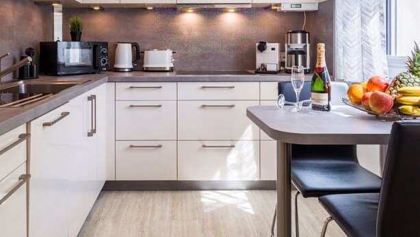 A modern kitchen with white cabinets, a stainless steel sink, a coffee maker, a microwave, and a toaster. There is a dining table with a chair, a bottle of champagne, and a fruit bowl. - Ferienwohnung Eden Apartments (Friedrichshafen)