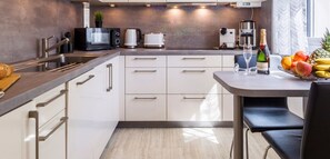 Eine moderne Küche mit weißen Schränken, einer Spüle aus Edelstahl, einer Kaffeemaschine, einer Mikrowelle und einem Toaster. Ein Esstisch mit einem Stuhl, eine Champagnerflasche und eine Obstschale sind ebenfalls vorhanden.