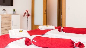 A hotel room with two beds, each with red towels and white linens. There is a wooden cabinet with a small plant and a sign on top. A television is mounted on the wall above the cabinet. - Ferienwohnung Eden Apartments (Friedrichshafen)