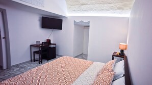 Ein kleiner, kompakter Raum mit einem Bett, einem Schreibtisch, einem Stuhl, einem an der Wand befestigten Fernseher und einer Lampe auf einem Nachttisch.
