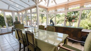 Un invernadero con una mesa y sillas de vidrio, rodeado de ventanales que dan a zonas verdes.