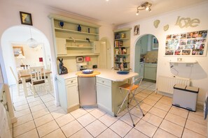 A compact kitchen with a dining area, featuring a small island, open shelving, and a wall-mounted photo collage. - 'Raggleswood' - a seaside home on Friars Cliff, Mudeford (Christchurch)