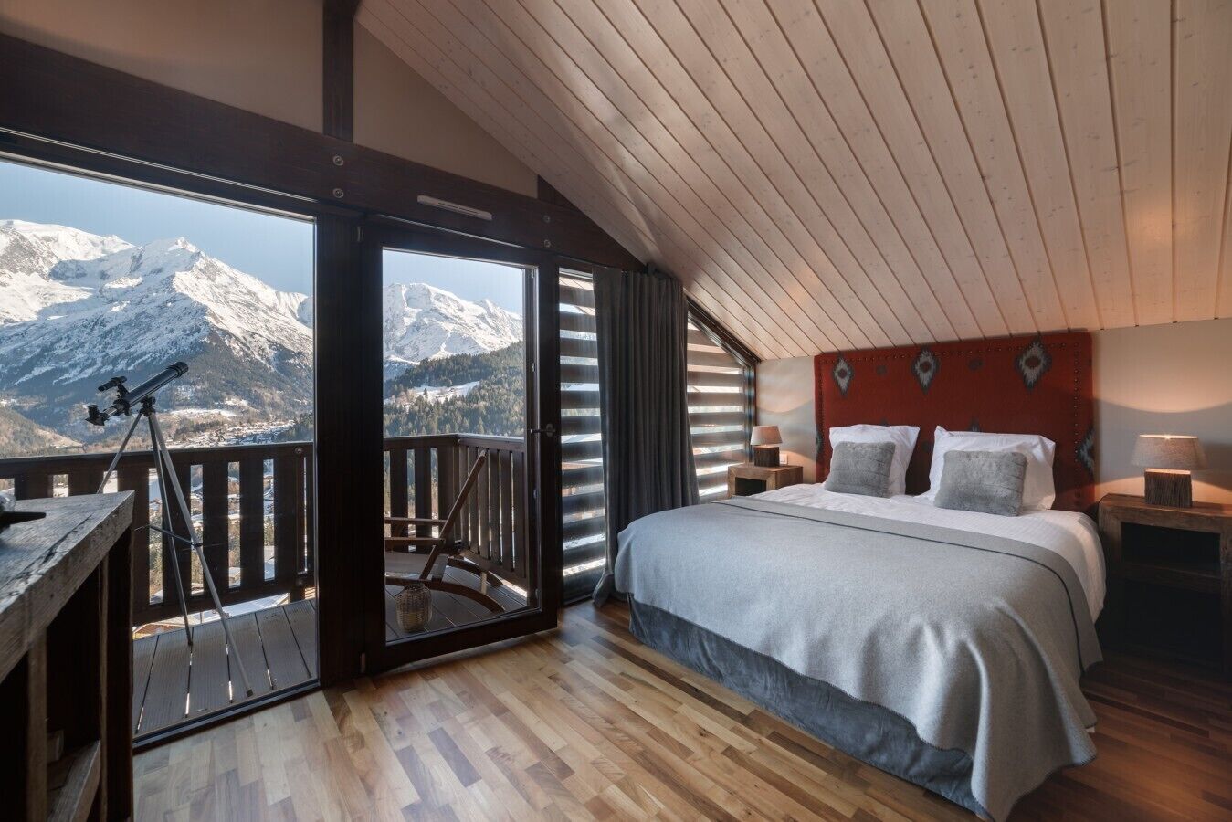 A bedroom with a bed, a nightstand, a lamp, a telescope on a balcony, and a view of snow-capped mountains.