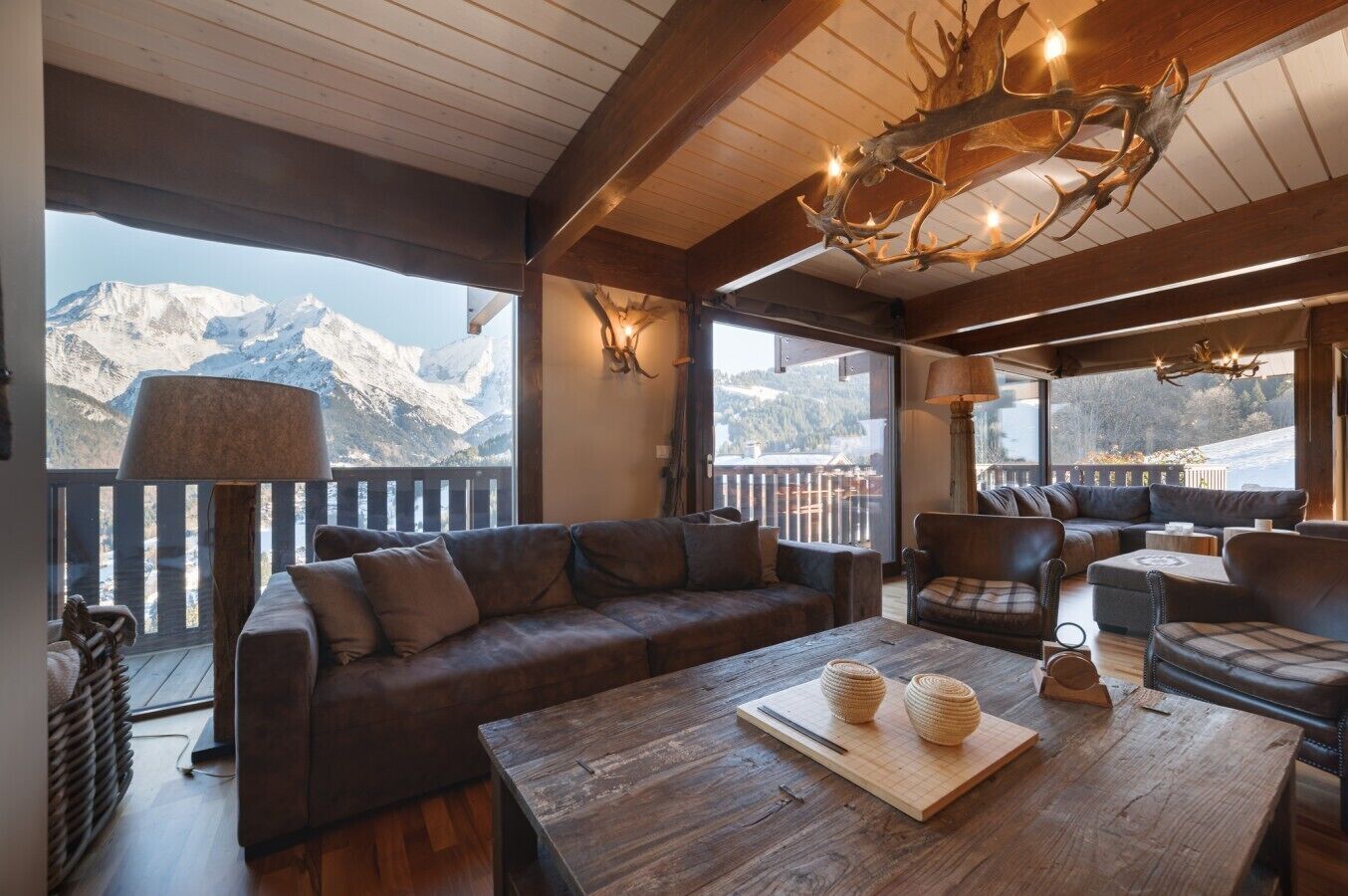 Ein gemütlicher Innenraum mit Holzbalkendecke, einem großen Fenster mit Bergblick, einer Couch, einem Tisch mit Dekoration und einer Lampe.
