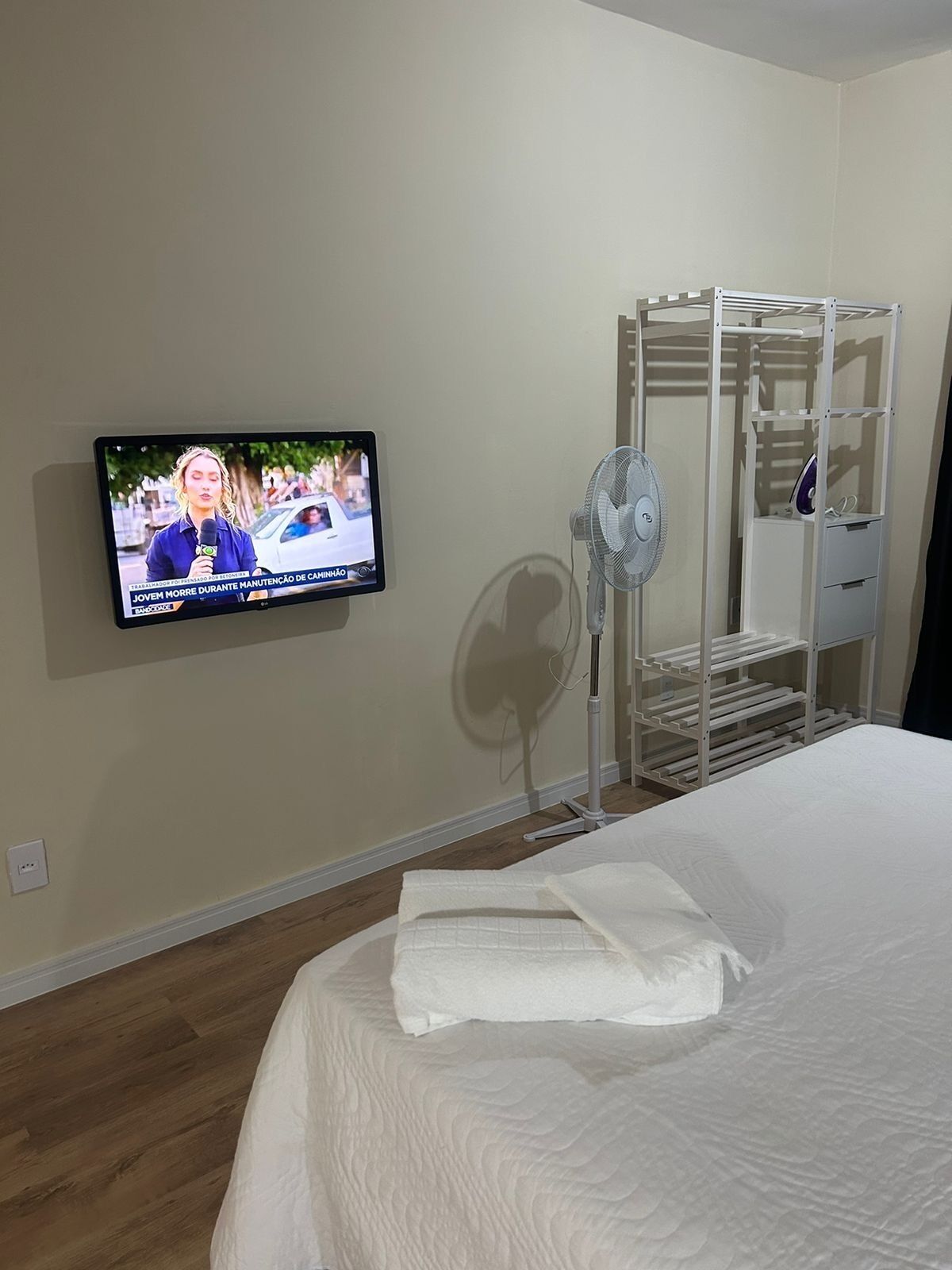 Habitación de hotel con cama, televisor montado en la pared, ventilador de pie y estantería metálica.