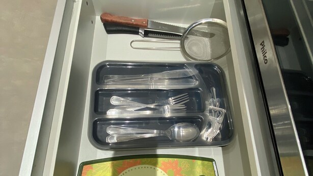 A drawer containing a knife, a pair of tongs, a spoon, and a cutting board with a floral design. - Smart Business Flat (Campinas)