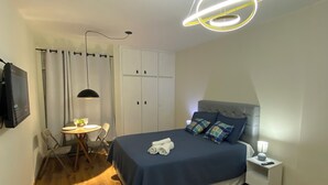 A bedroom with a bed, bedside table, lamp, and a dining area with chairs and a table. - Smart Business Flat (Campinas)