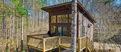 A wooden cabin with a deck and a staircase, surrounded by trees.