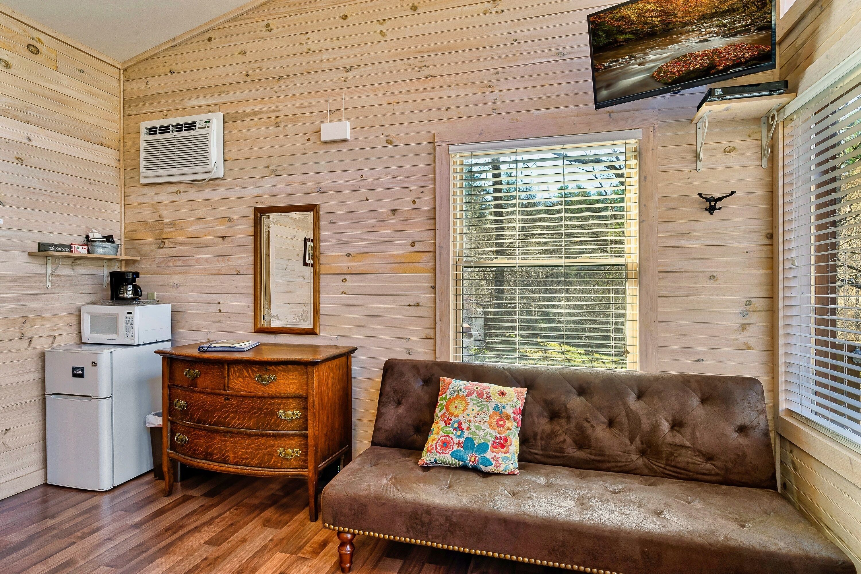 Una habitación acogedora con paredes de madera, un sofá marrón con un cojín floral, un aparador de madera, un refrigerador blanco, un microondas y un televisor en la pared.