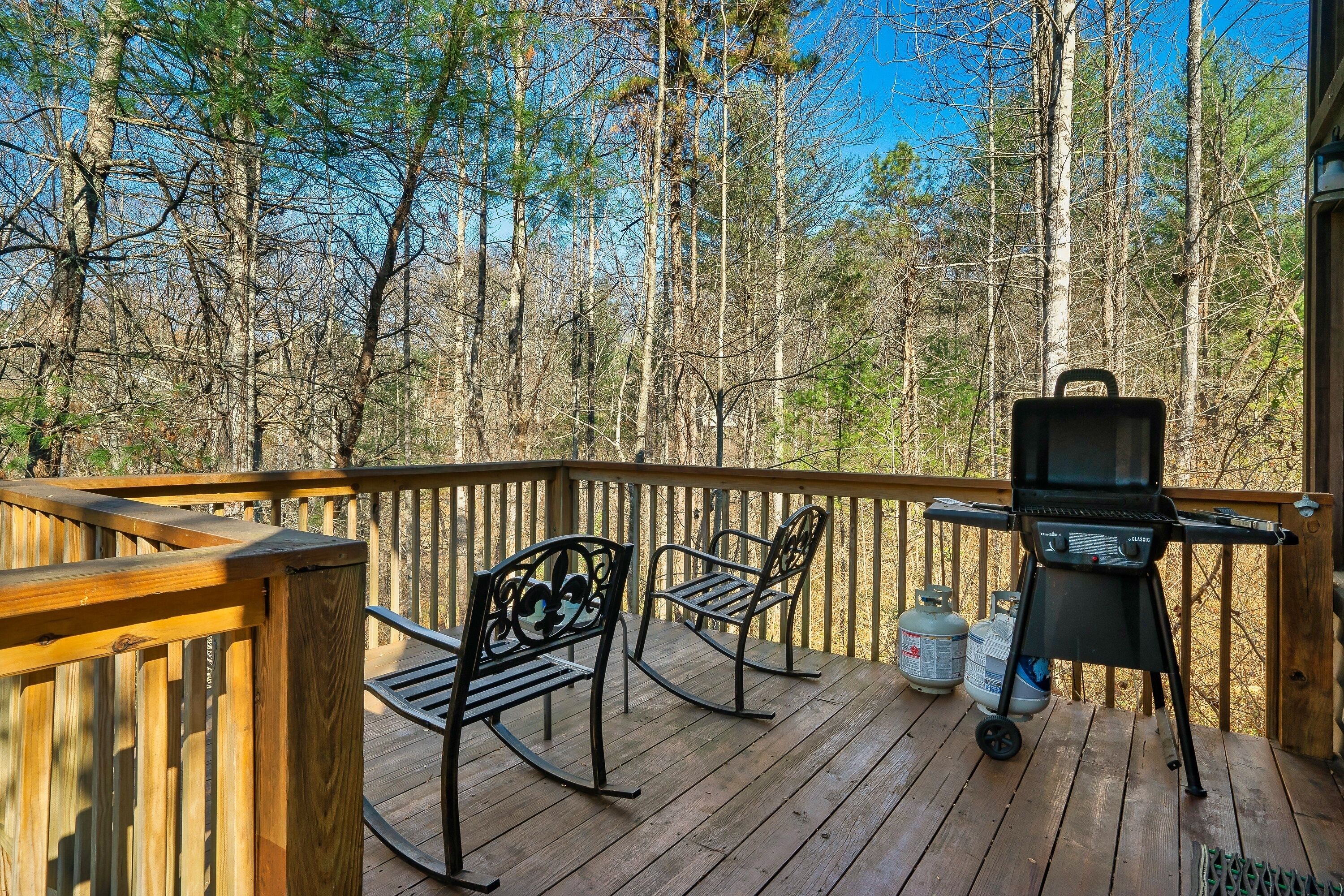 Eine Holzterrasse mit Grill und zwei Schaukelstühlen, umgeben von Bäumen.