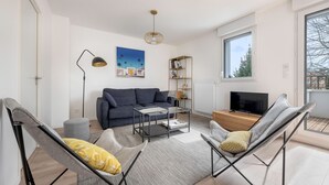 Ein modernes Wohnzimmer mit einer Couch, zwei Sesseln, einem Couchtisch, einem Fernseher, einem Bücherregal und einem Fenster mit Blick.