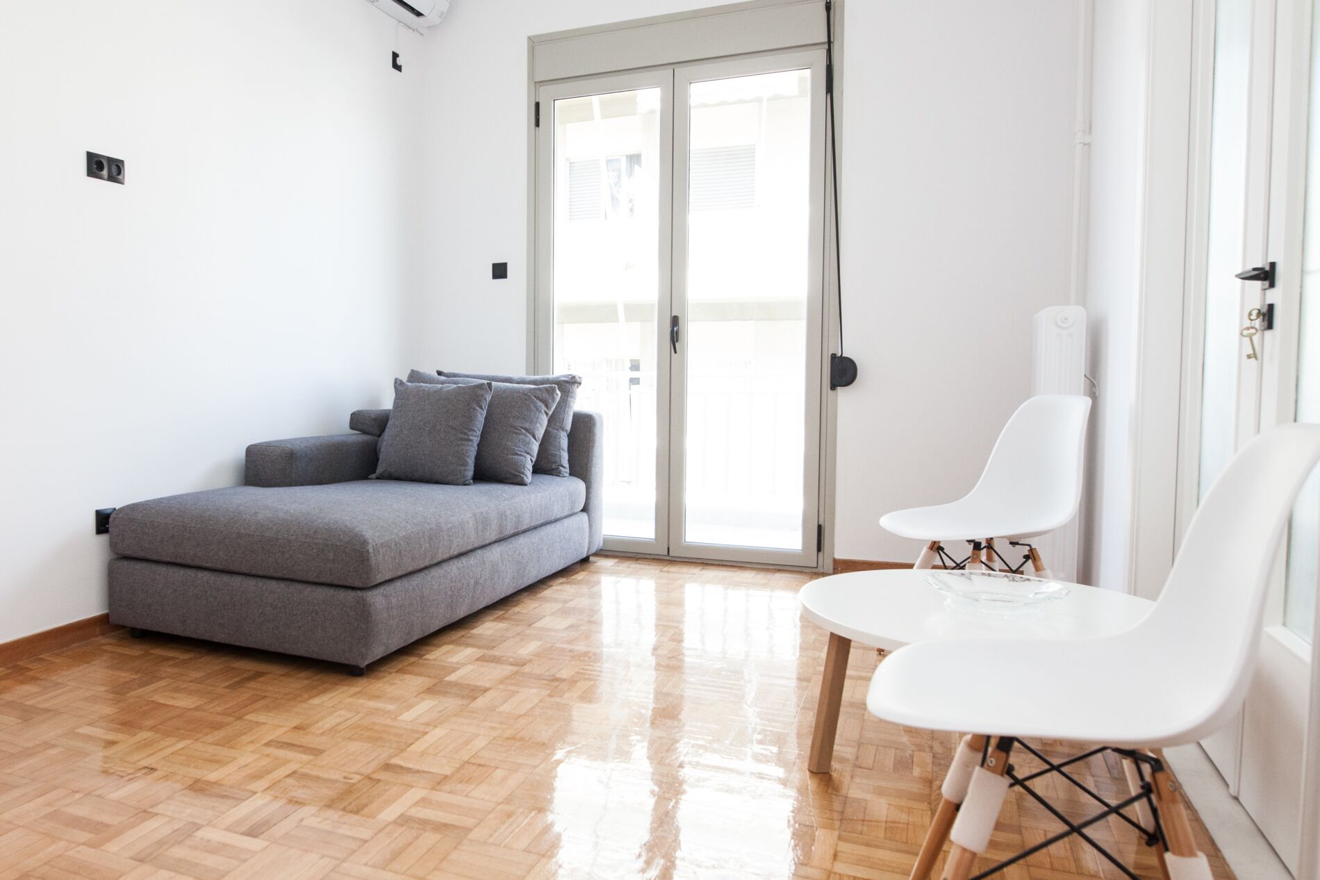 Un salon moderne avec un canapé gris, deux fauteuils blancs, un parquet en bois et une porte-fenêtre coulissante donnant sur un espace extérieur.
