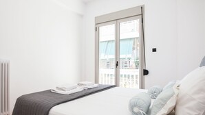 Un dormitorio con cama, radiador, ventana con vista a edificios y puerta.