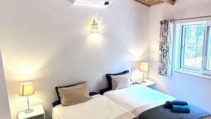 A room with a thatched ceiling, two beds with dark and light bedding, a bedside table with a lamp, and a window with curtains. - Vues Spectaculaires sur L'océan.fibre (BORDEIRA)