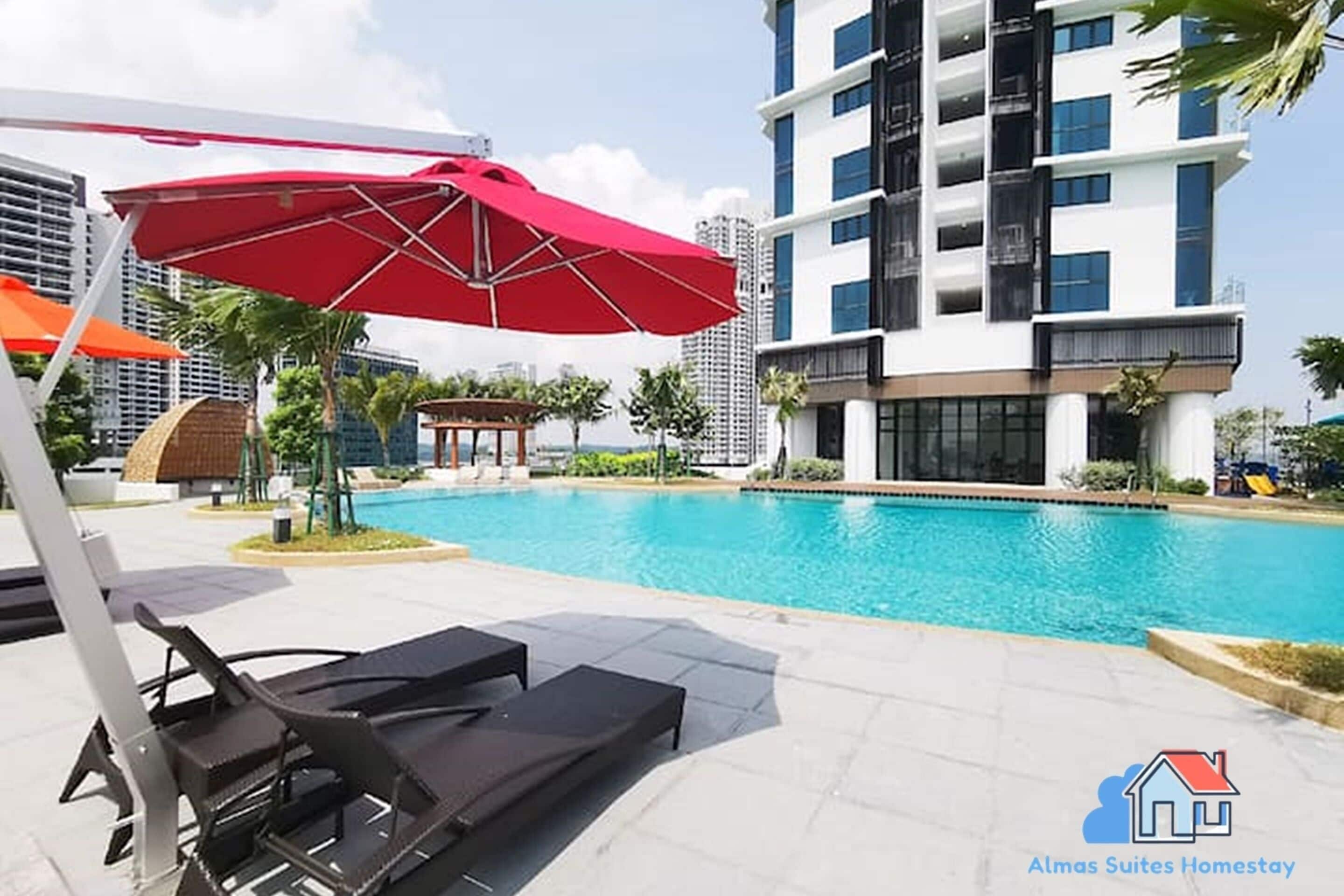 A poolside area with lounge chairs and a red umbrella, surrounded by modern buildings and a clear sky.