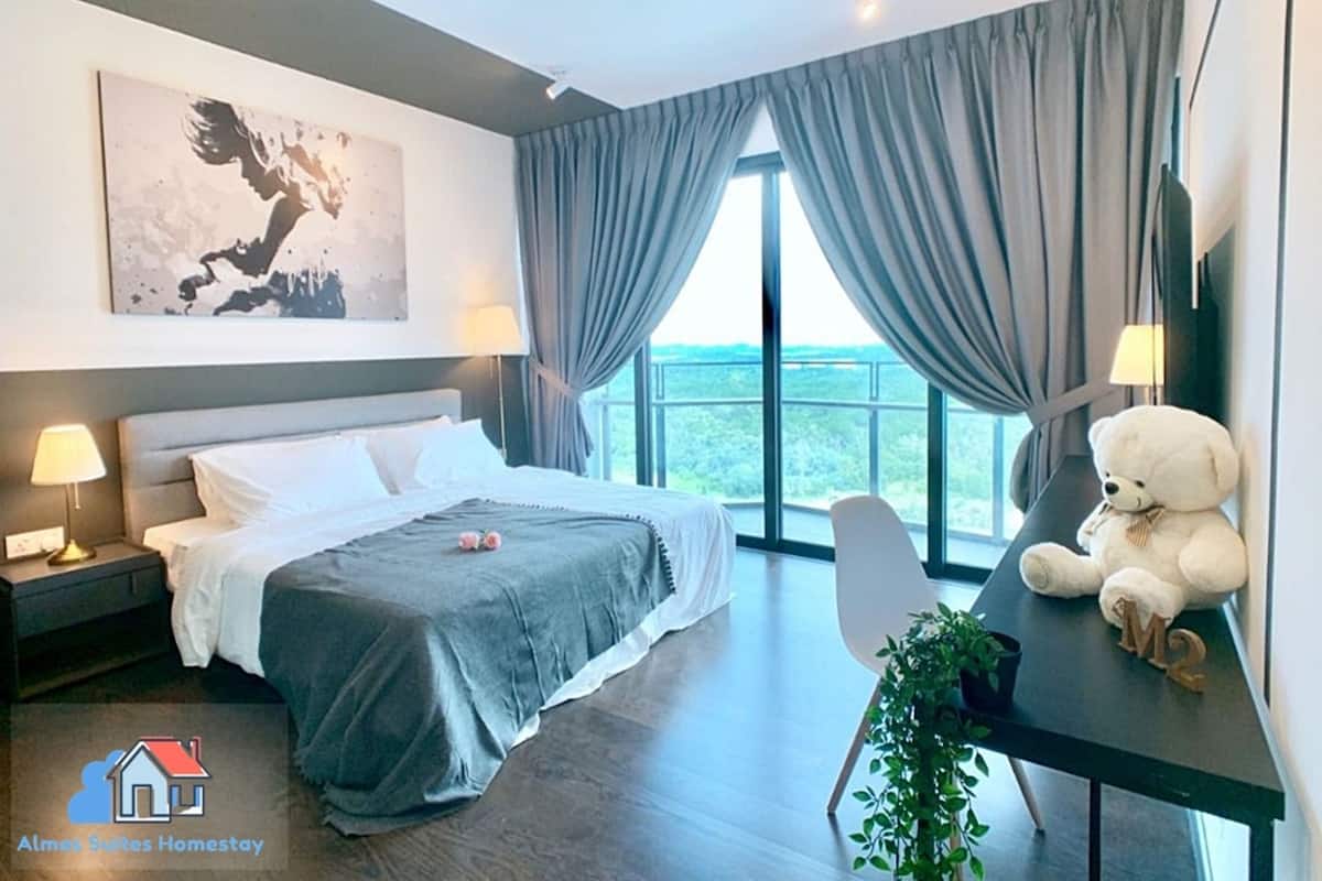 A modern hotel room with a bed, bedside lamps, a desk with a teddy bear, a potted plant, and a large window with curtains.