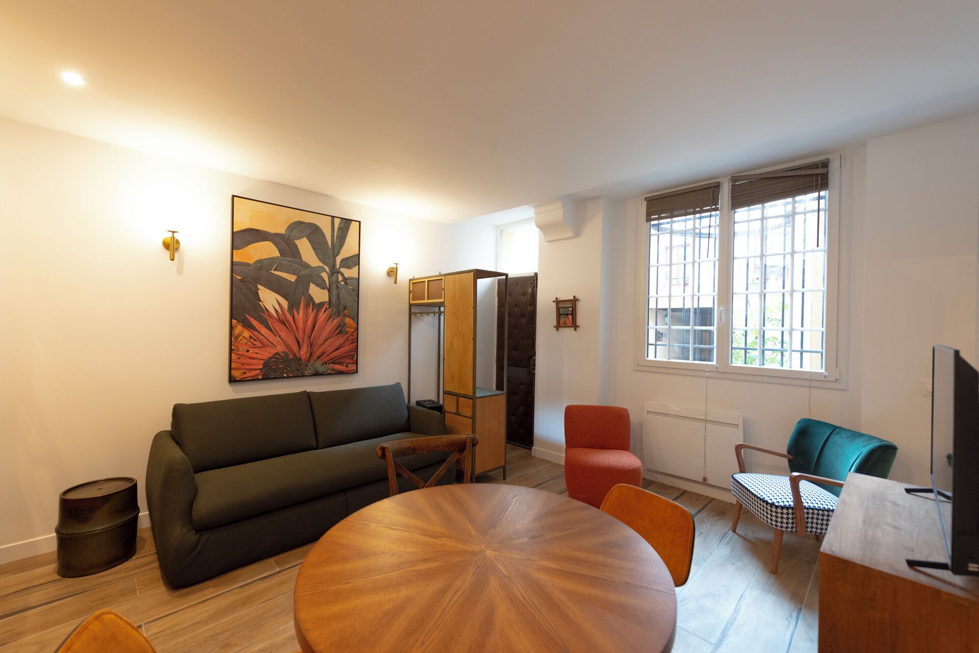 A modern living room with a sofa, a round wooden table, and a red armchair. There is a large window with blinds, a painting on the wall, and a television on a stand.