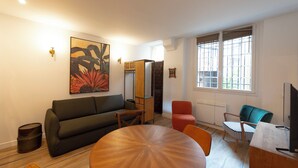 A modern living room with a sofa, a round wooden table, and a red armchair. There is a large window with blinds, a painting on the wall, and a television on a stand. - Au pied de la Tour, 1BR/1BA, 4 people (París)