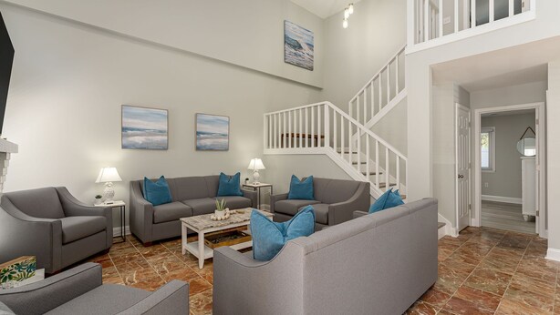 A spacious living room with grey sofas and blue cushions, a coffee table, and two wall-mounted paintings. A white staircase leads to an upper level. - Pet Friendly, Recently Renovated, Private Hot Tub + Free Attraction Tickets! (North Myrtle Beach)