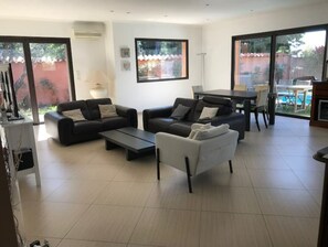 A modern living room with two sofas, a coffee table, and a dining area with a table and chairs. Large windows provide views of the outdoors.