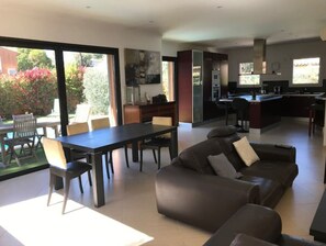 A modern living room with a dining area, a kitchen, and a large sliding glass door leading to a garden.
