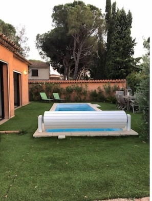 A backyard with a swimming pool, a lawn, and a patio area with chairs.