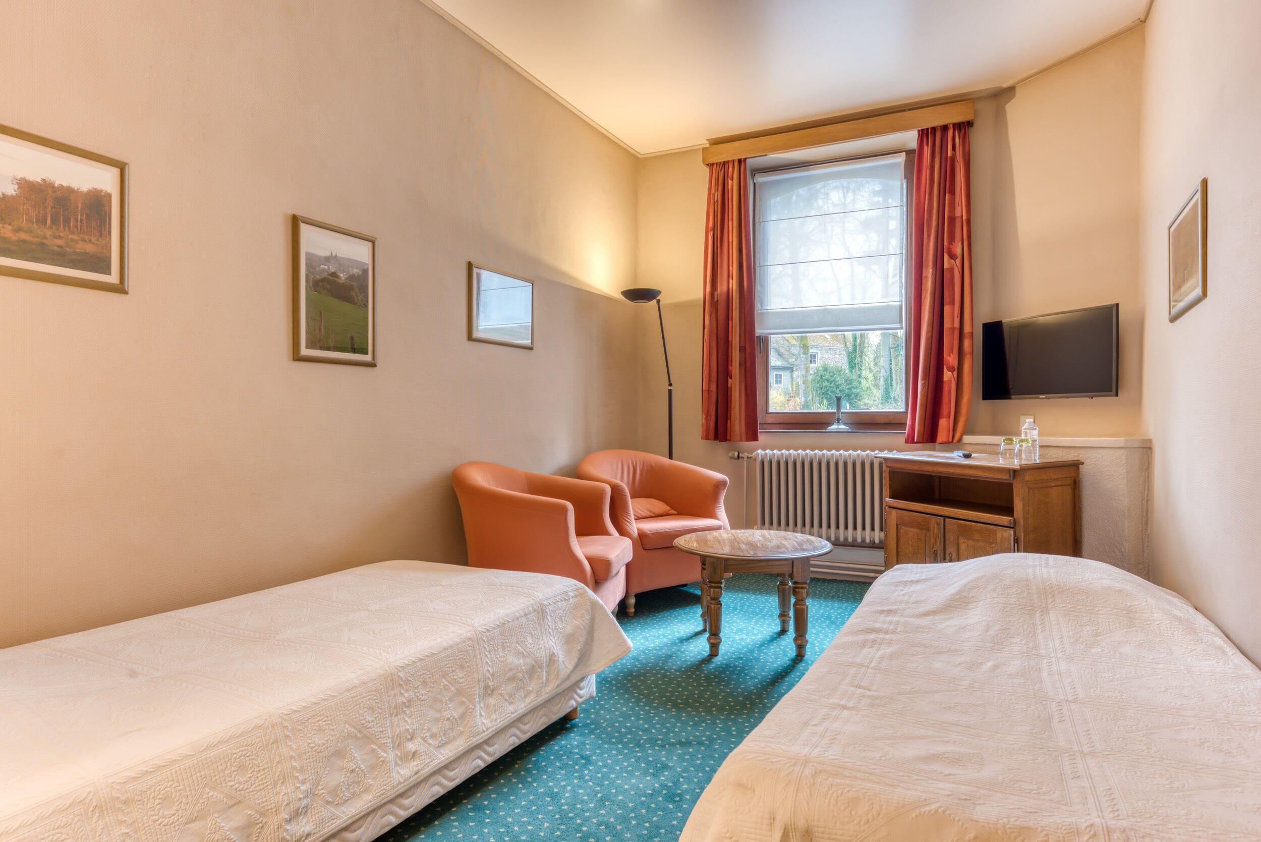 A hotel room with two single beds, a desk with a computer, a chair, a radiator, and a window with curtains.