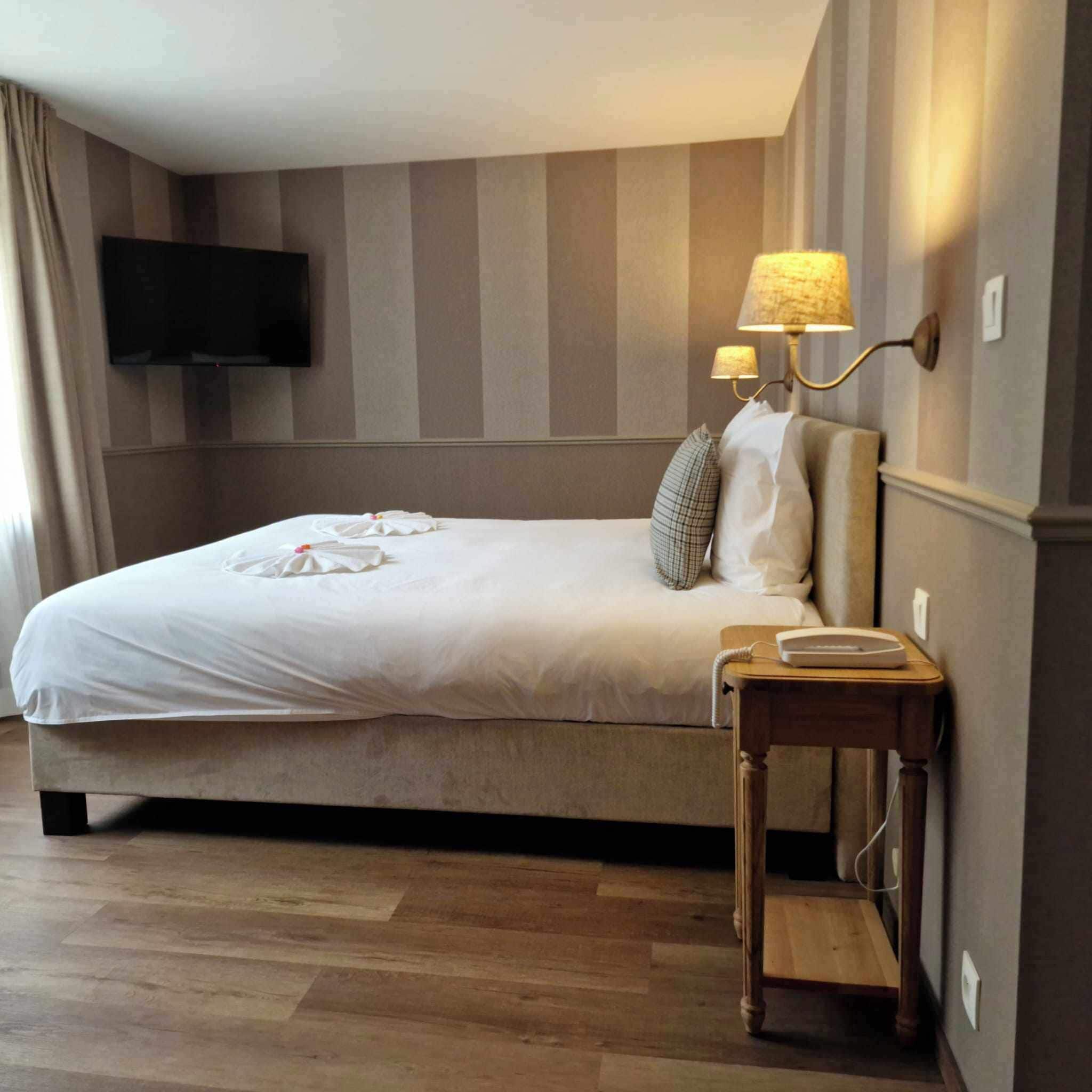 A hotel room with a large bed, a bedside table with a lamp, a flat-screen TV mounted on the wall, and a wooden floor.