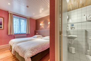 A bedroom with two beds, a window with curtains, a radiator, and a bathroom with a sink and mirror. - Hôtel Le Cor de Chasse (Saint-Hubert)