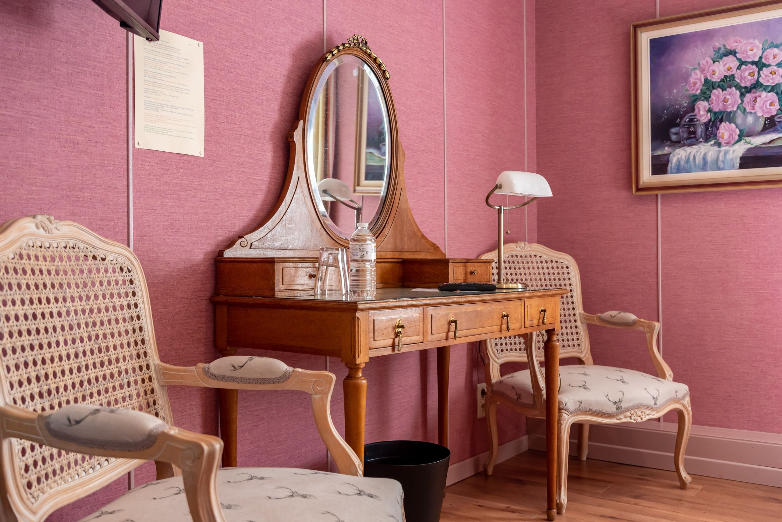 A room with a wooden vanity, two ornate chairs, a mirror, a lamp, and a framed floral painting on the wall.