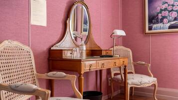 A room with a wooden vanity, two ornate chairs, a mirror, a lamp, and a framed floral painting on the wall.