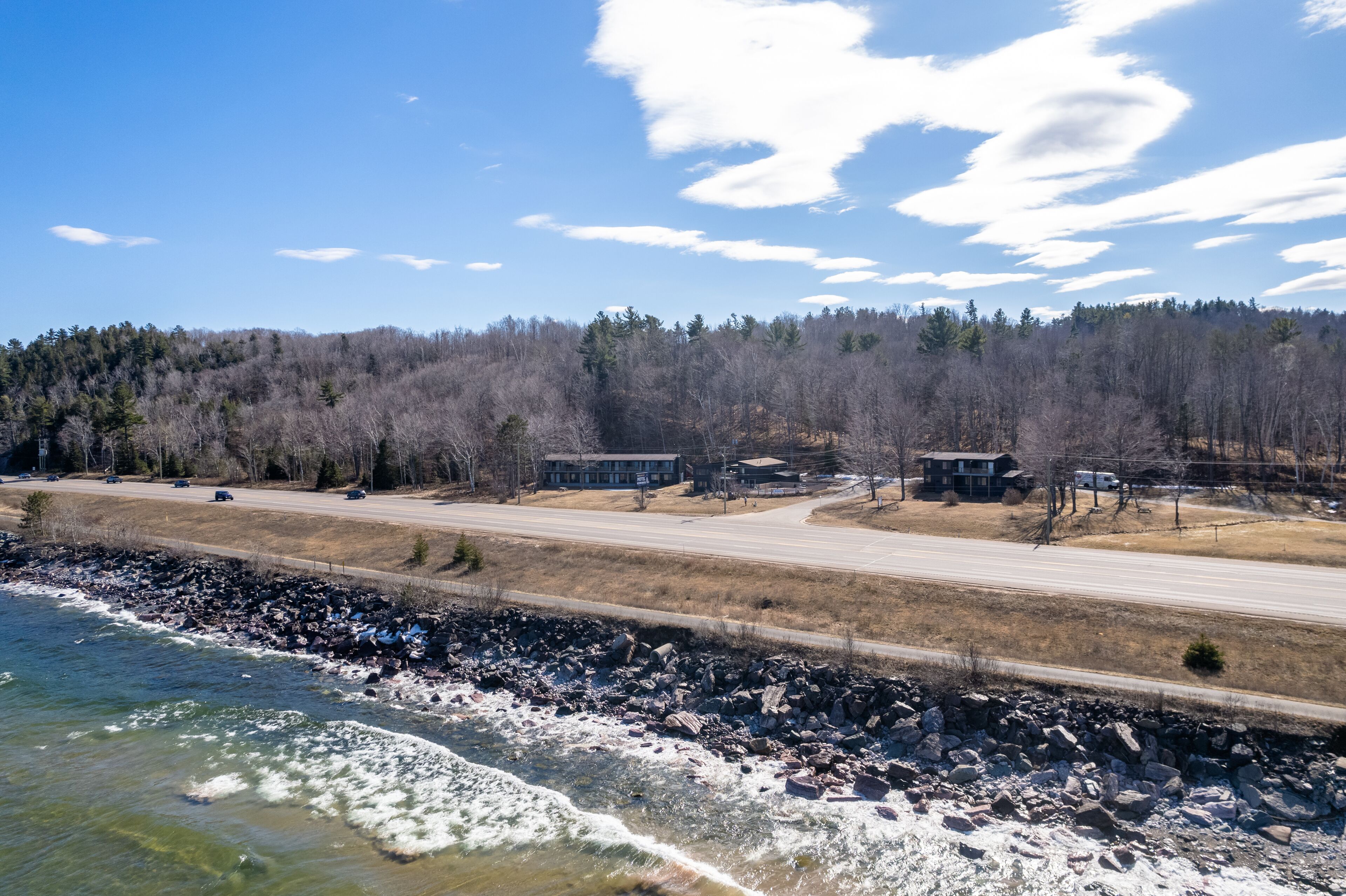 A coastal road with a rocky shoreline, a forested hill in the background, and a clear sky.