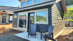 Una casa moderna de dos pisos con un amplio ventanal corredero, un patio con dos sillas Adirondack y un letrero de dirección visible.