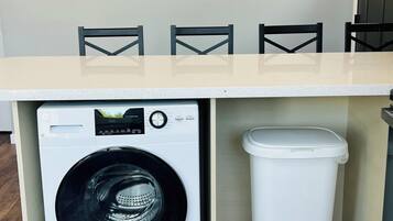 A compact laundry area with a washing machine, a trash bin, and a table with chairs.